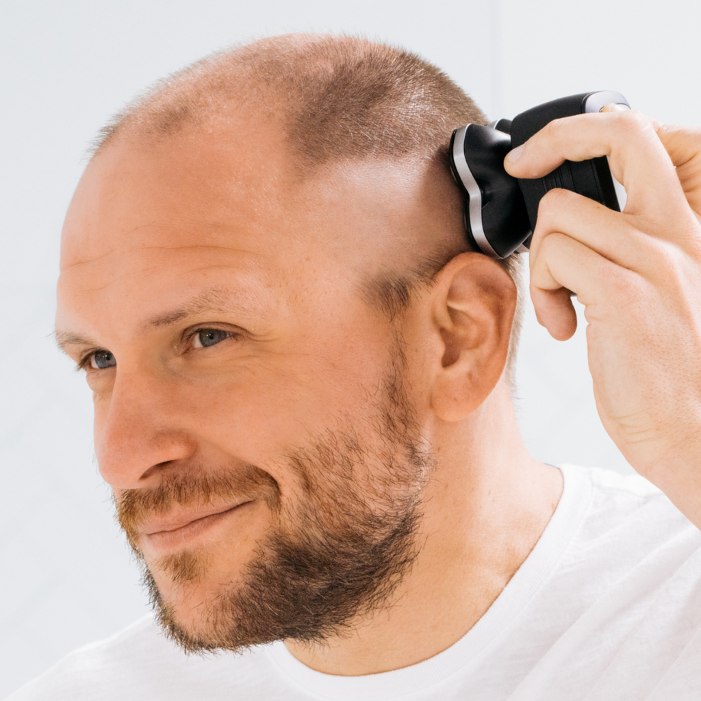Man using an electric razor to shave his head, smiling slightly, wearing a white t-shirt.
